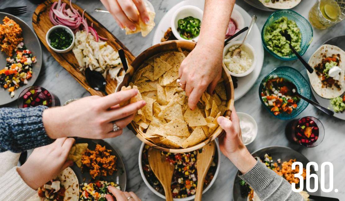 Manos compartiendo comida al estilo de un antiguo simposio: disfrutar primero, cuestionar después.